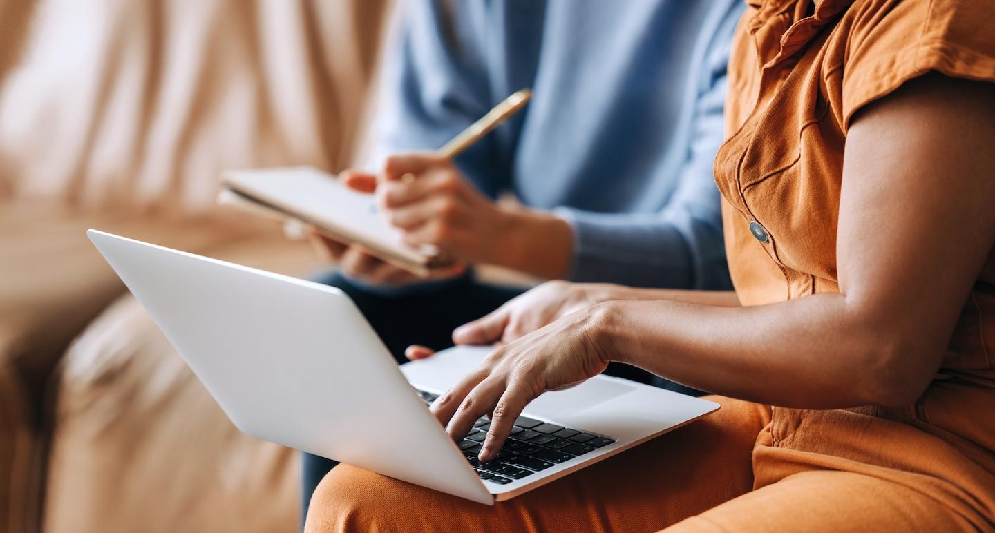 Closeup shot of a person in an office working on a laptop and another in the background taking notes on a notepad.