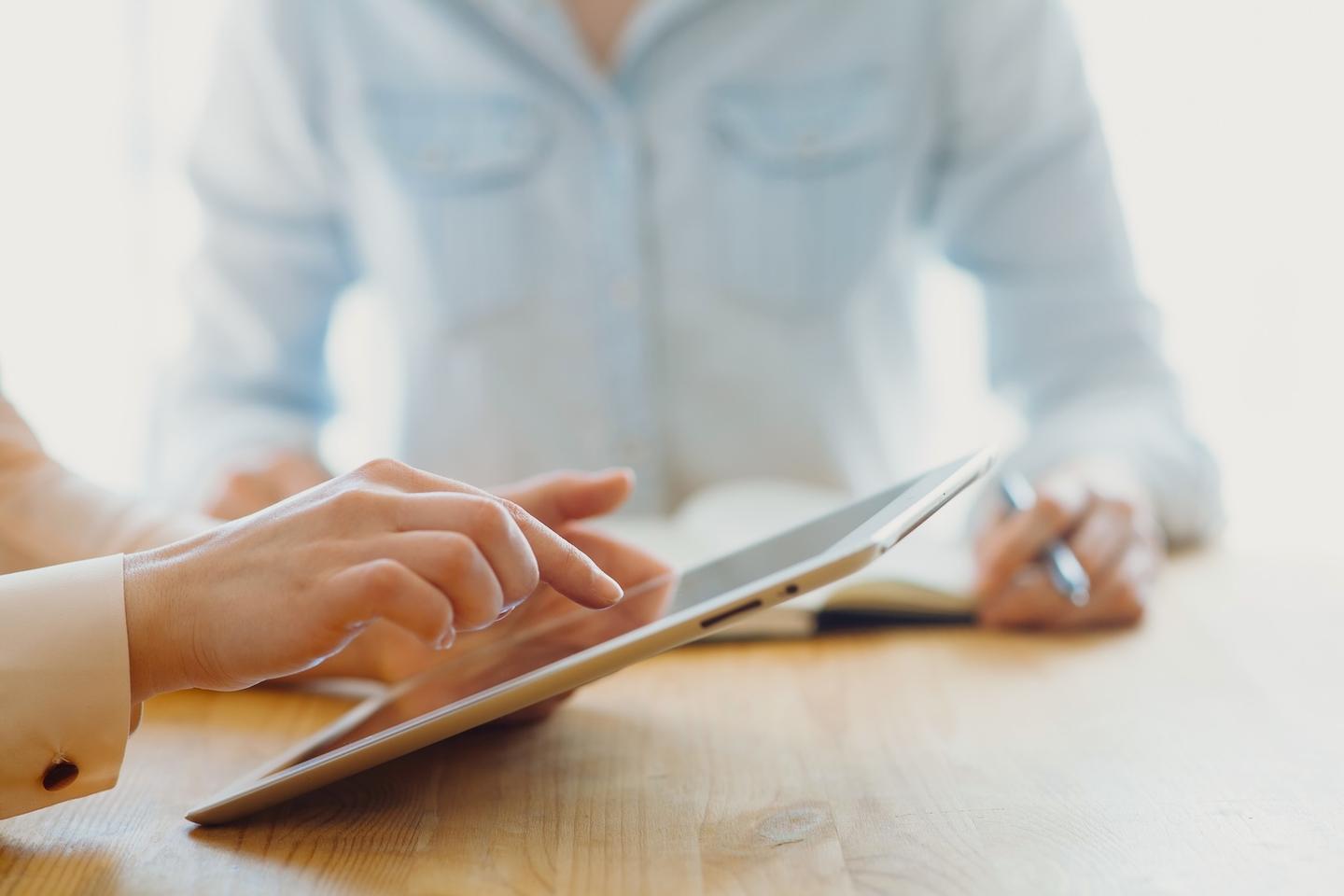 A person working on an iPad at a wooden table and another in the background writing in a book.