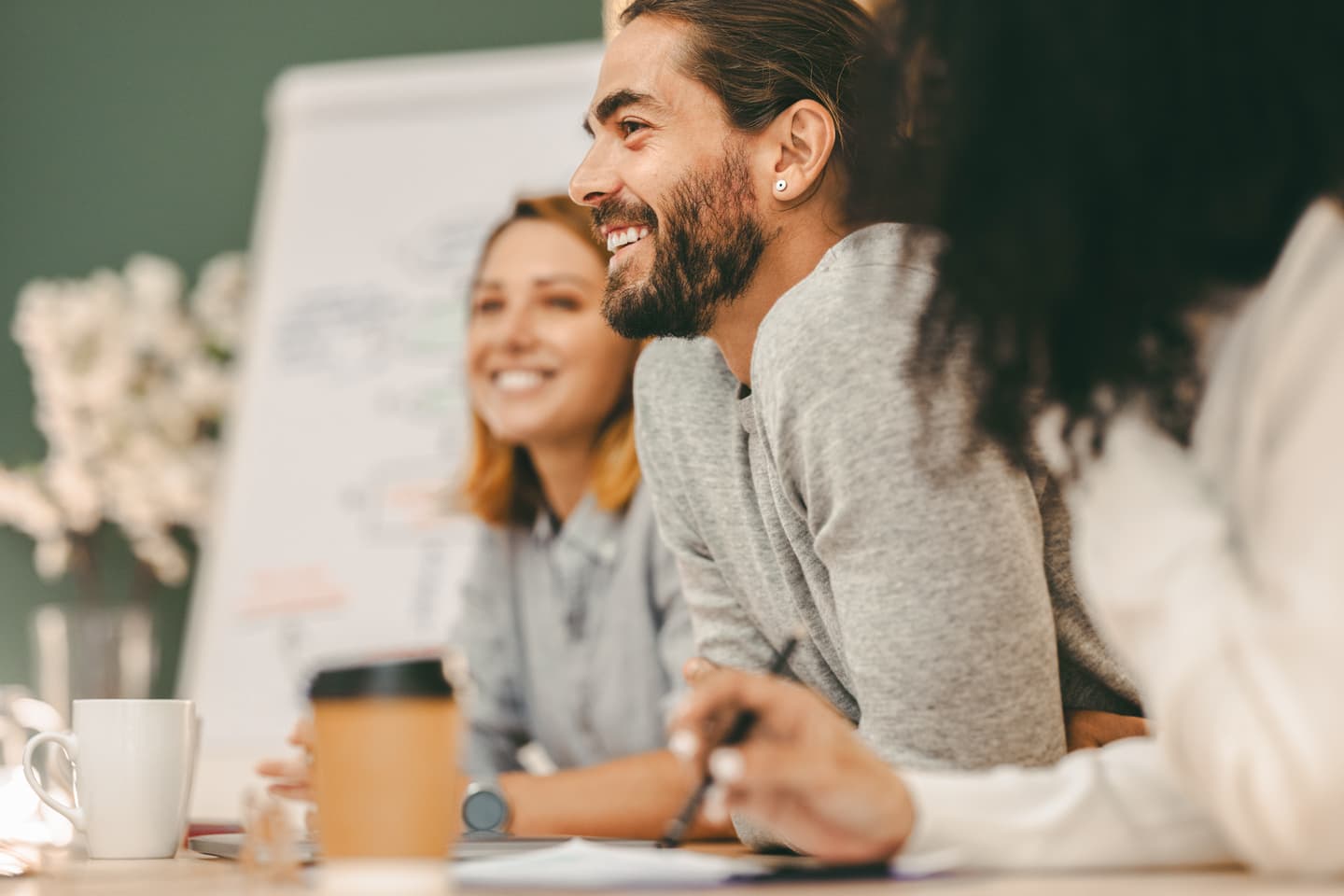Smiling people in a meeting with coffee cups on table, man with beard and bun in grey jumper in foreground.