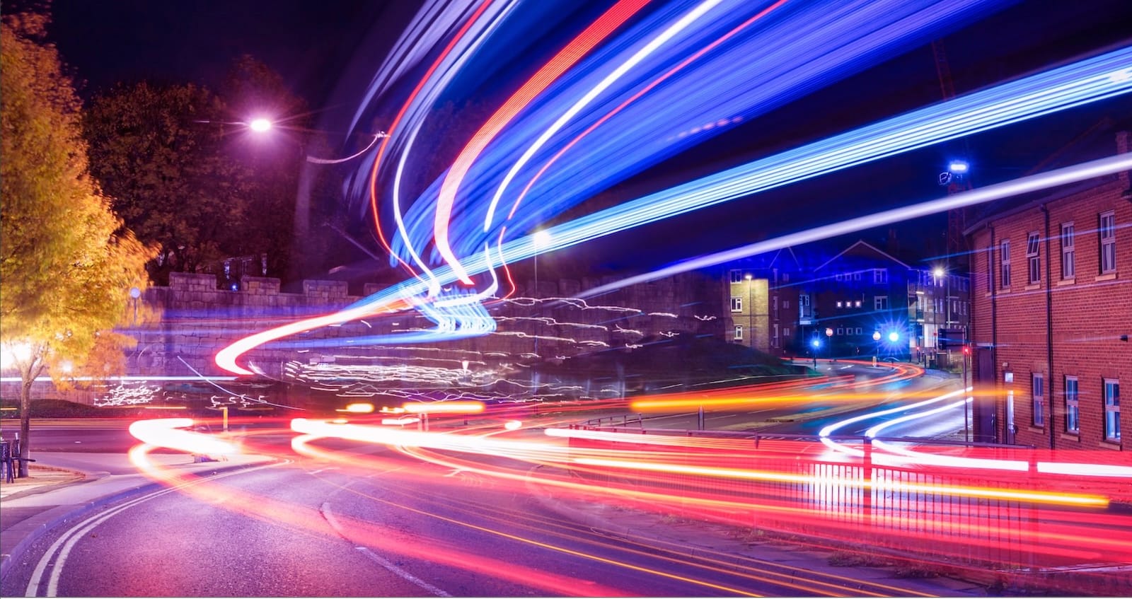 Long exposure night photograph showing colourful light trails from traffic against urban buildings and autumn trees.