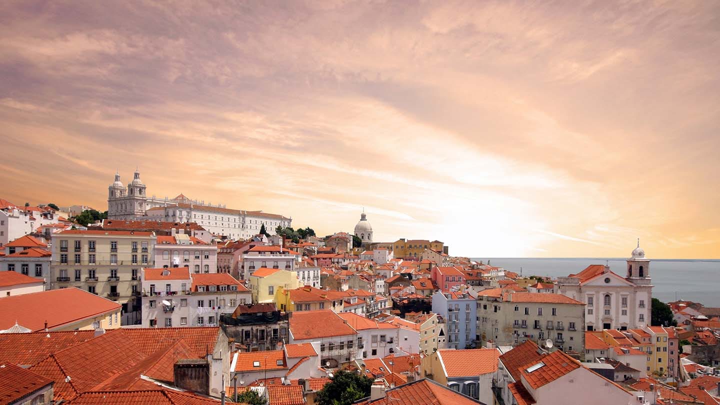 Lisbon cityscape with orange terracotta rooftops, historic buildings and cathedral under a golden sunset sky.