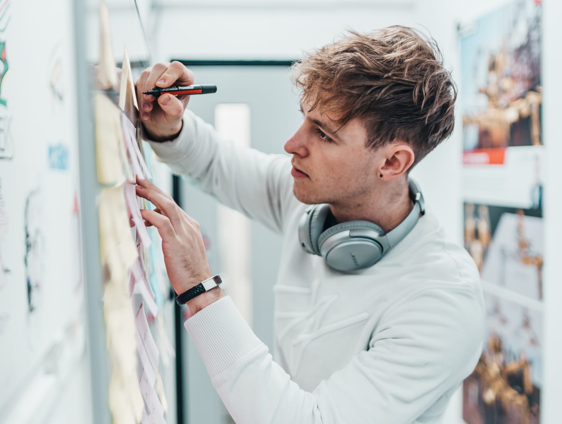 Person in white jumper with headphones writing on wall notes, focused on creative planning in bright studio space.