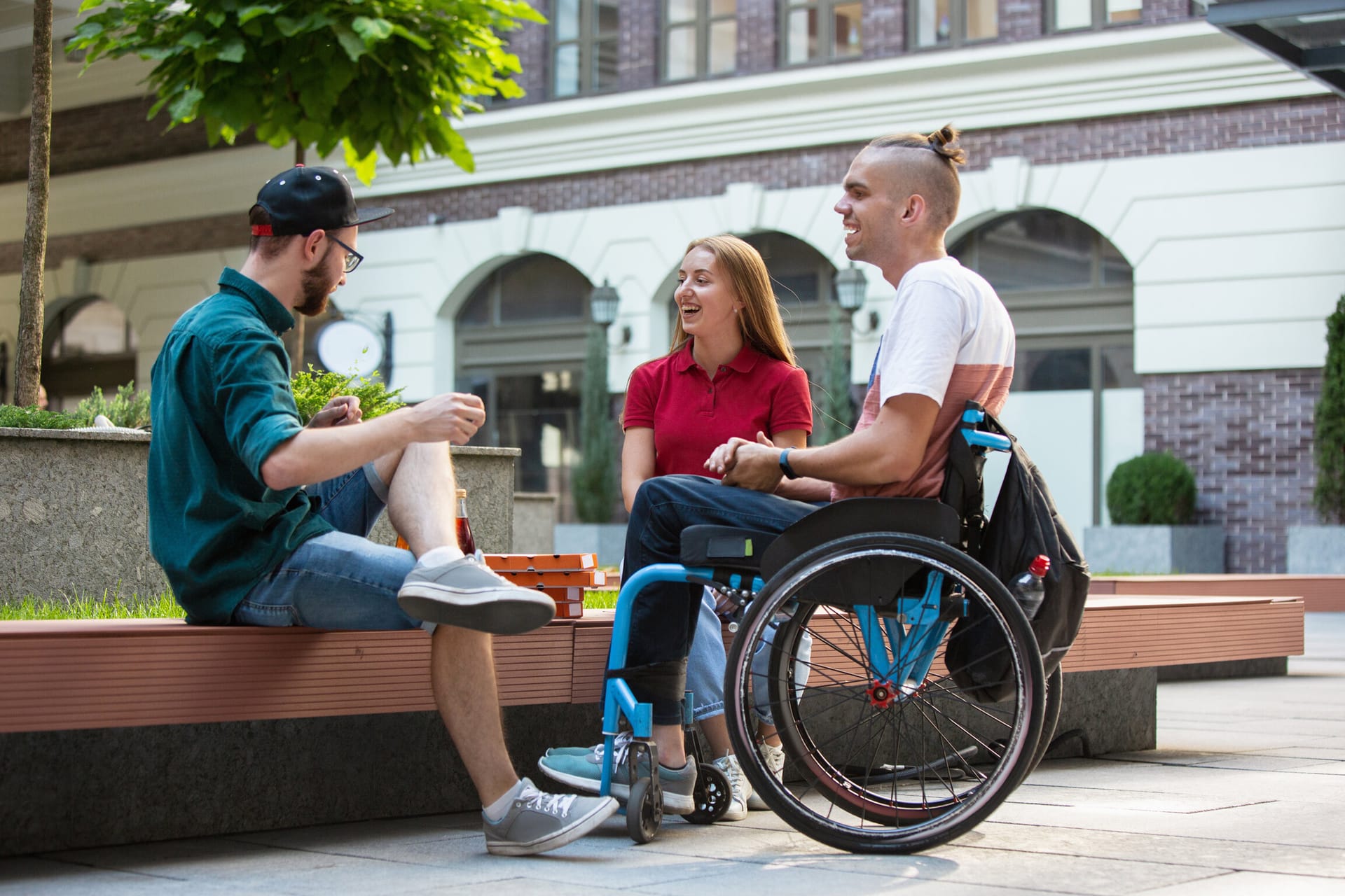 Three friends chatting outdoors, one using a wheelchair, sitting by a bench near a brick building with greenery.