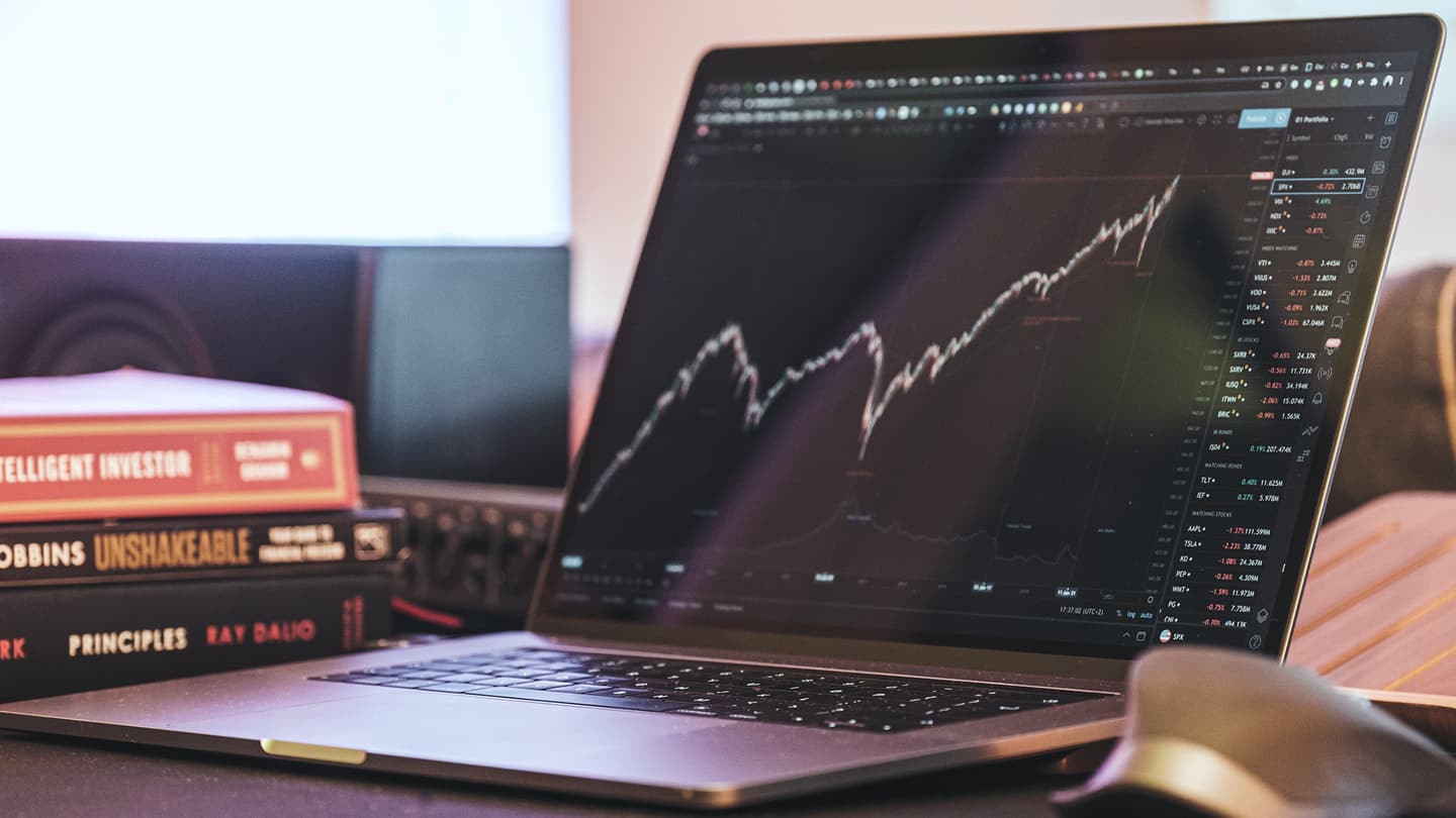 A laptop displaying stock market data and graphs, next to books about investing.