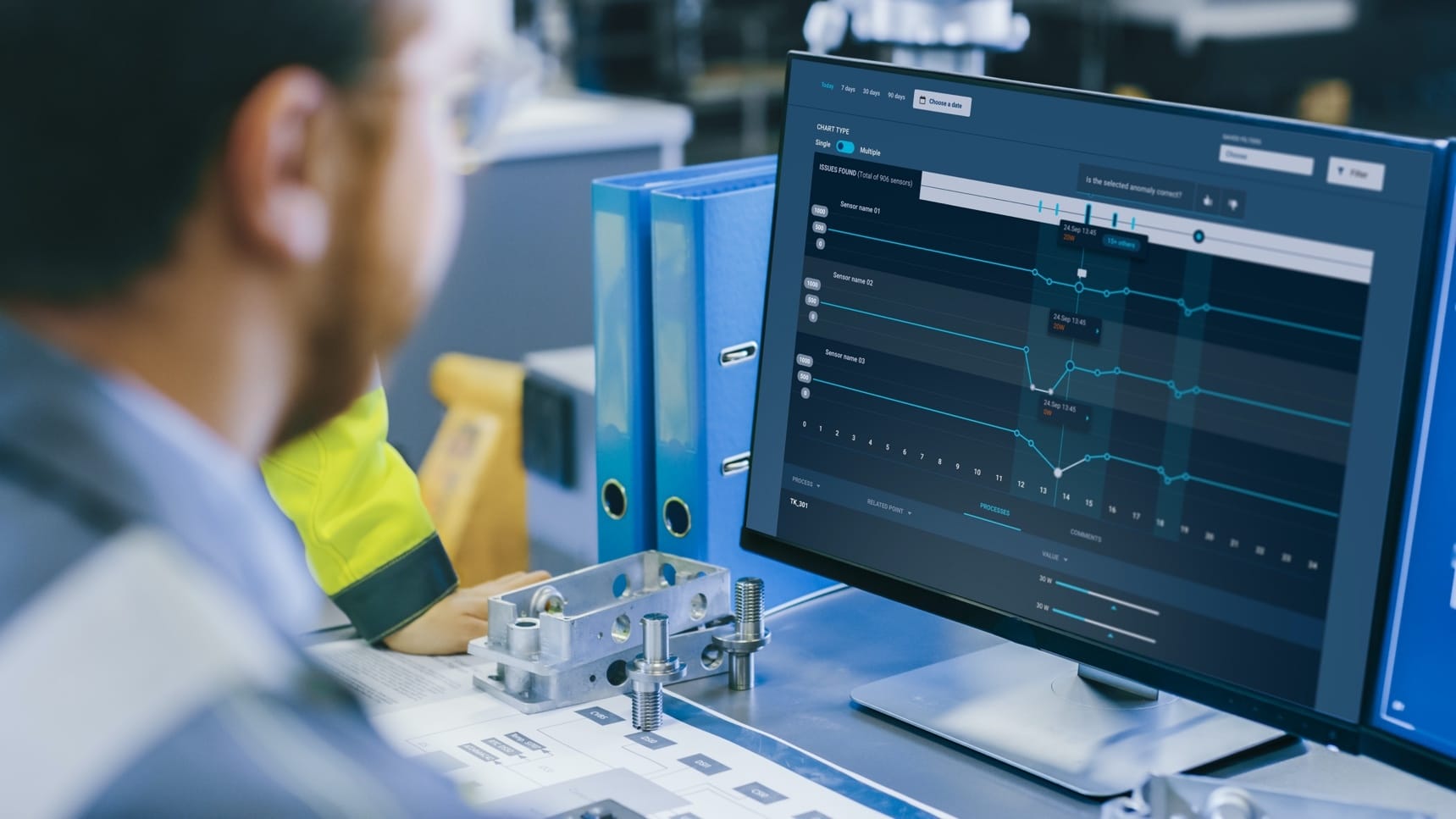 Person viewing data analytics dashboard with blue line graphs on computer screen, metal components and blue folders nearby.