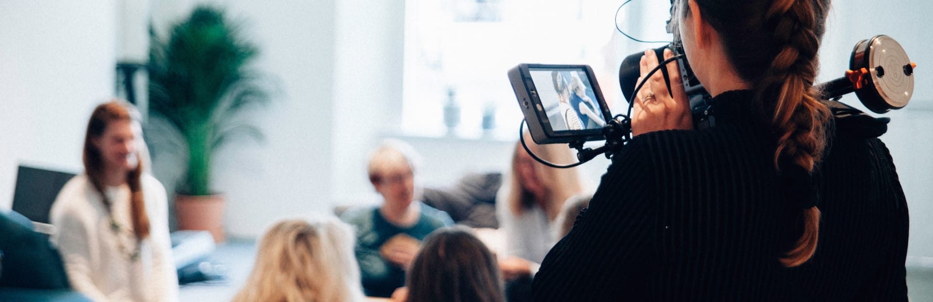 Person filming a group discussion with a camera in a bright office space with a green plant in the background.