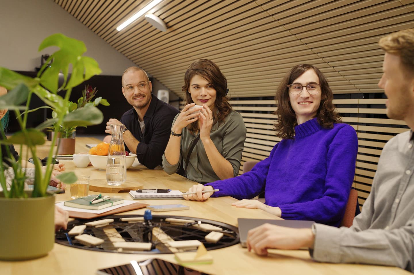 Group meeting around wooden table with refreshments in modern office space with wooden slat ceiling and green plant.
