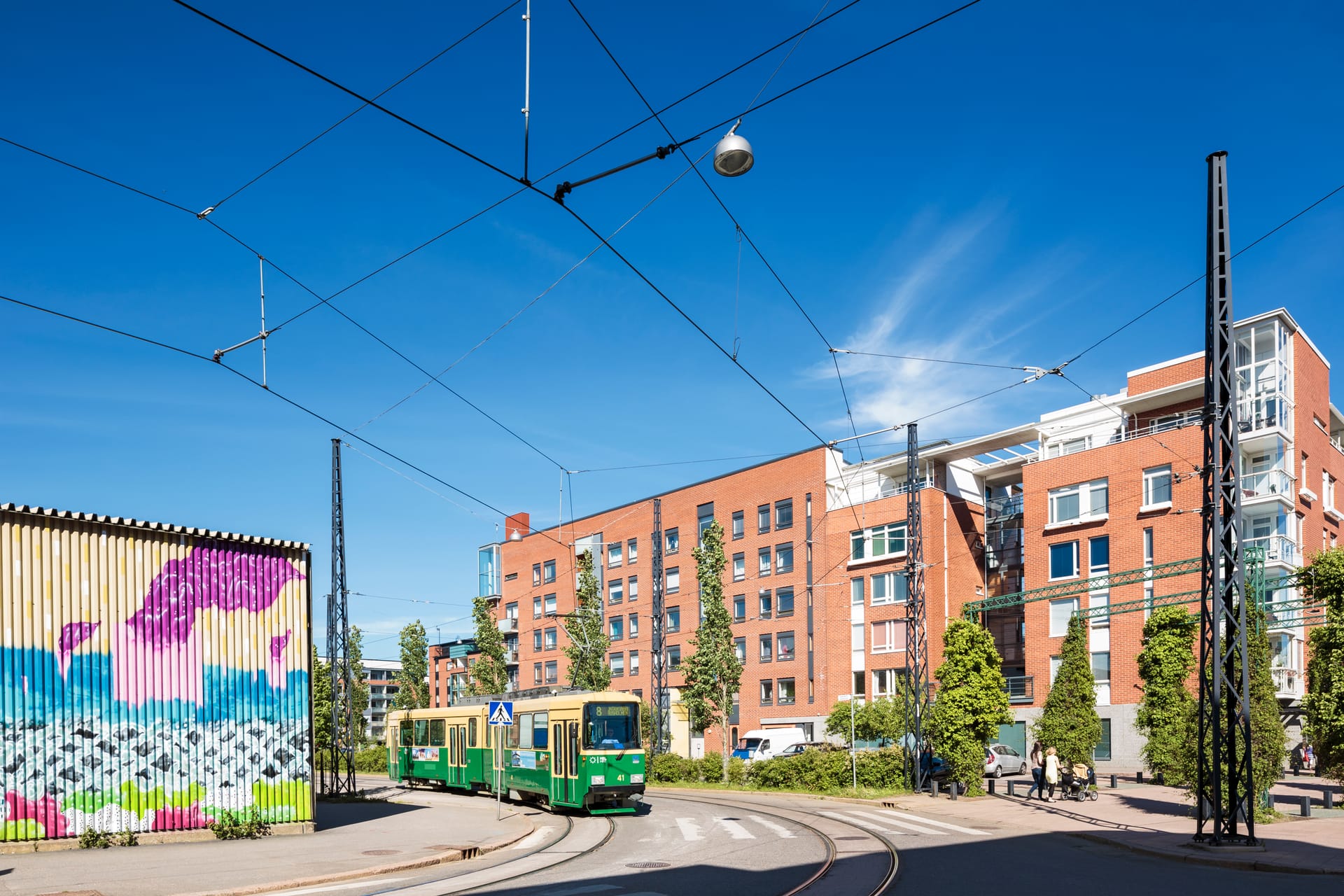 Green tram on tracks in urban setting with brick apartment buildings, colourful mural, and overhead power lines.
