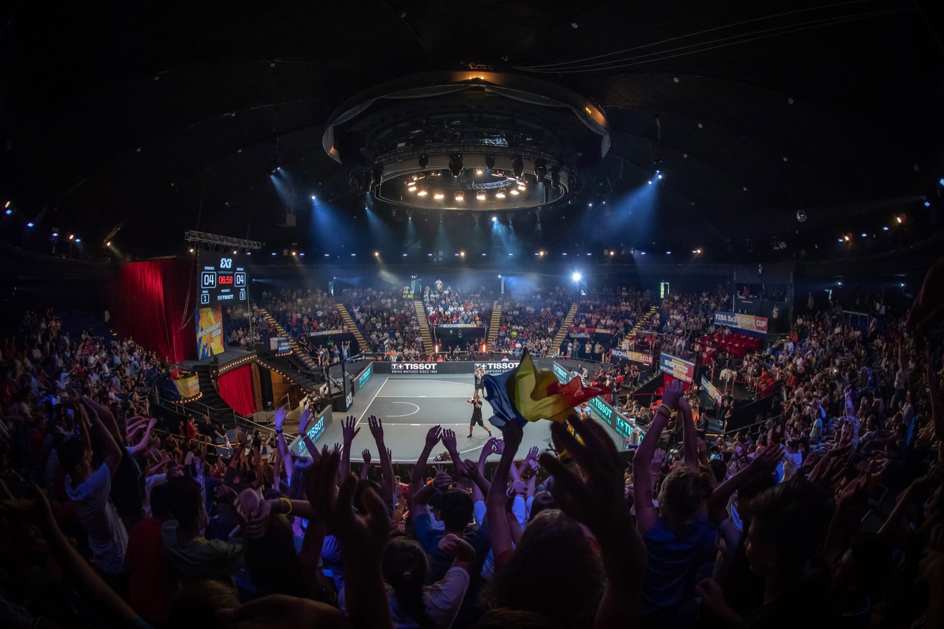 Indoor basketball arena with enthusiastic crowd watching a FIBA 3x3 tournament, dramatic lighting and scoreboard visible.