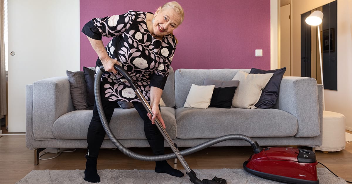 Smiling person in floral dress vacuuming with red Miele hoover beside grey sofa in room with purple wall.