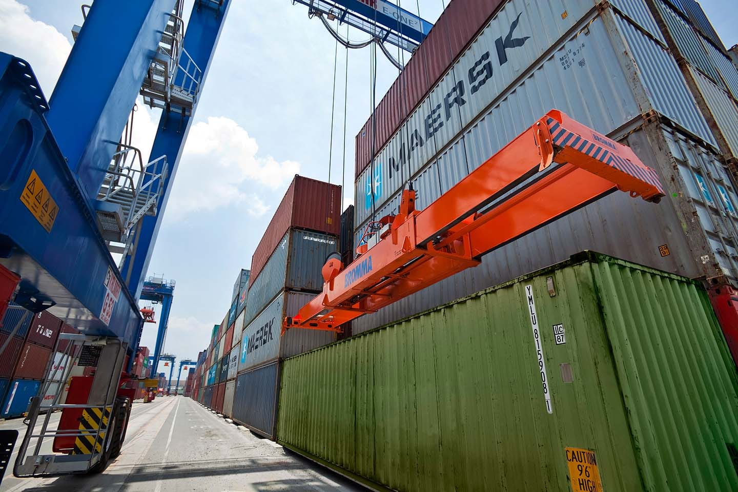 Shipping containers stacked at port terminal with blue crane and orange lifting equipment handling Maersk containers.