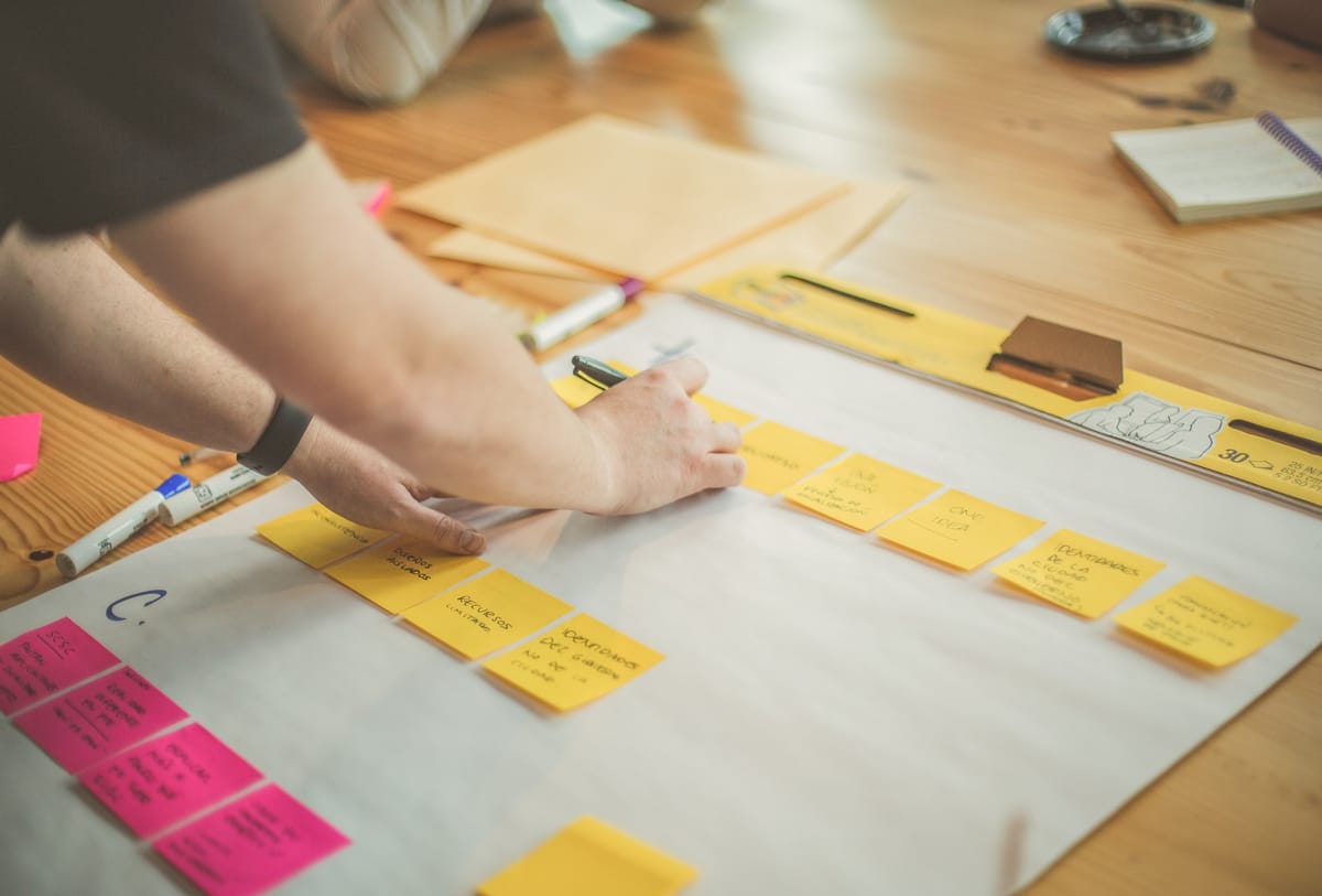 Person arranging yellow and pink sticky notes on a paper during a planning or brainstorming session on wooden desk.