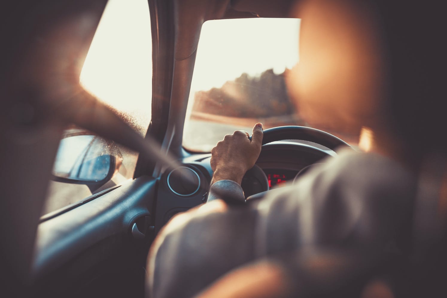Person driving a car during sunset, with hand on steering wheel and warm golden light streaming through the window.