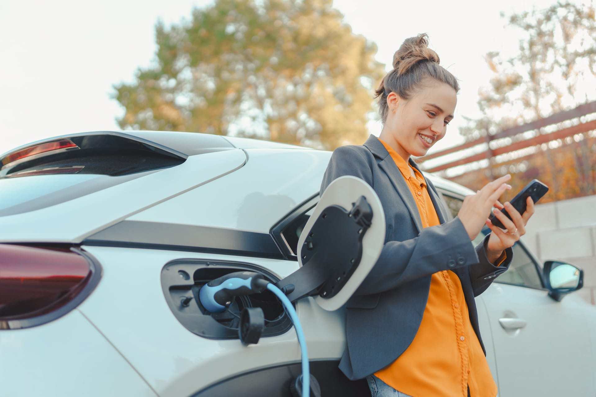 Person in blue blazer and orange shirt checking phone while charging white electric car outdoors in an autumn setting.