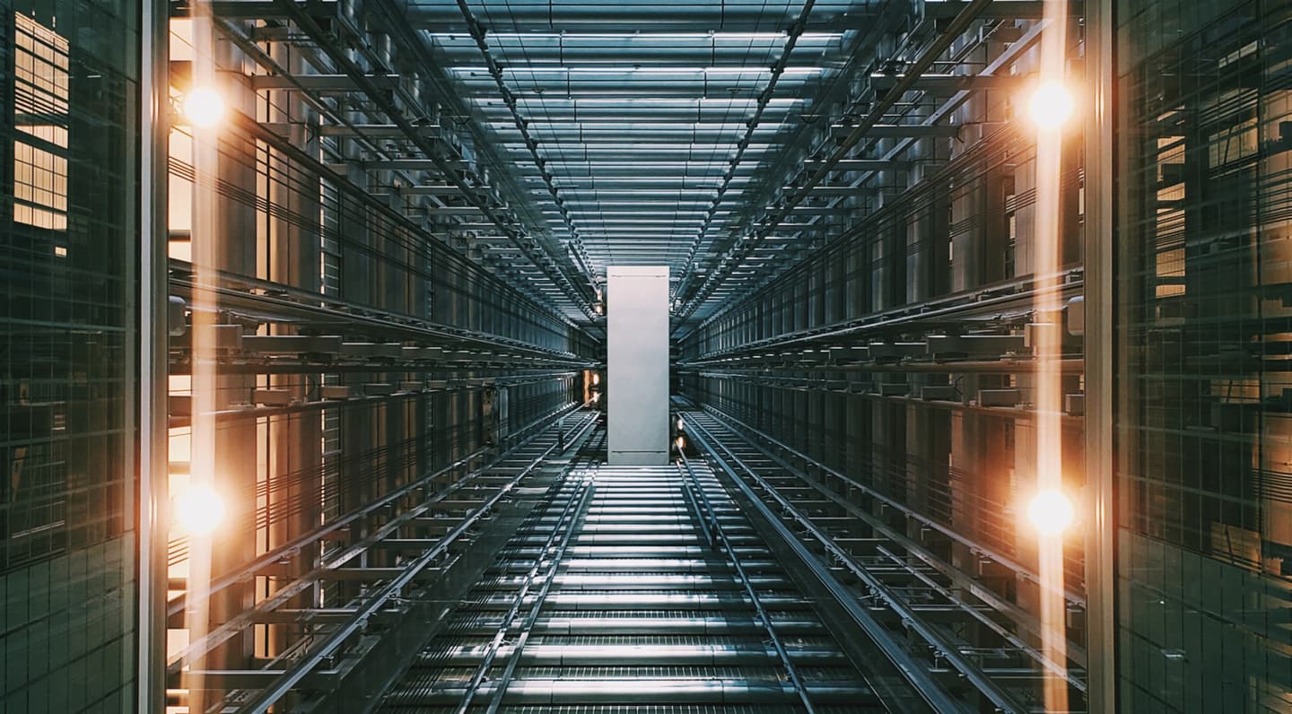 Looking up through a modern elevator shaft with glass walls, illuminated by warm lights, with a lift car visible at the top.