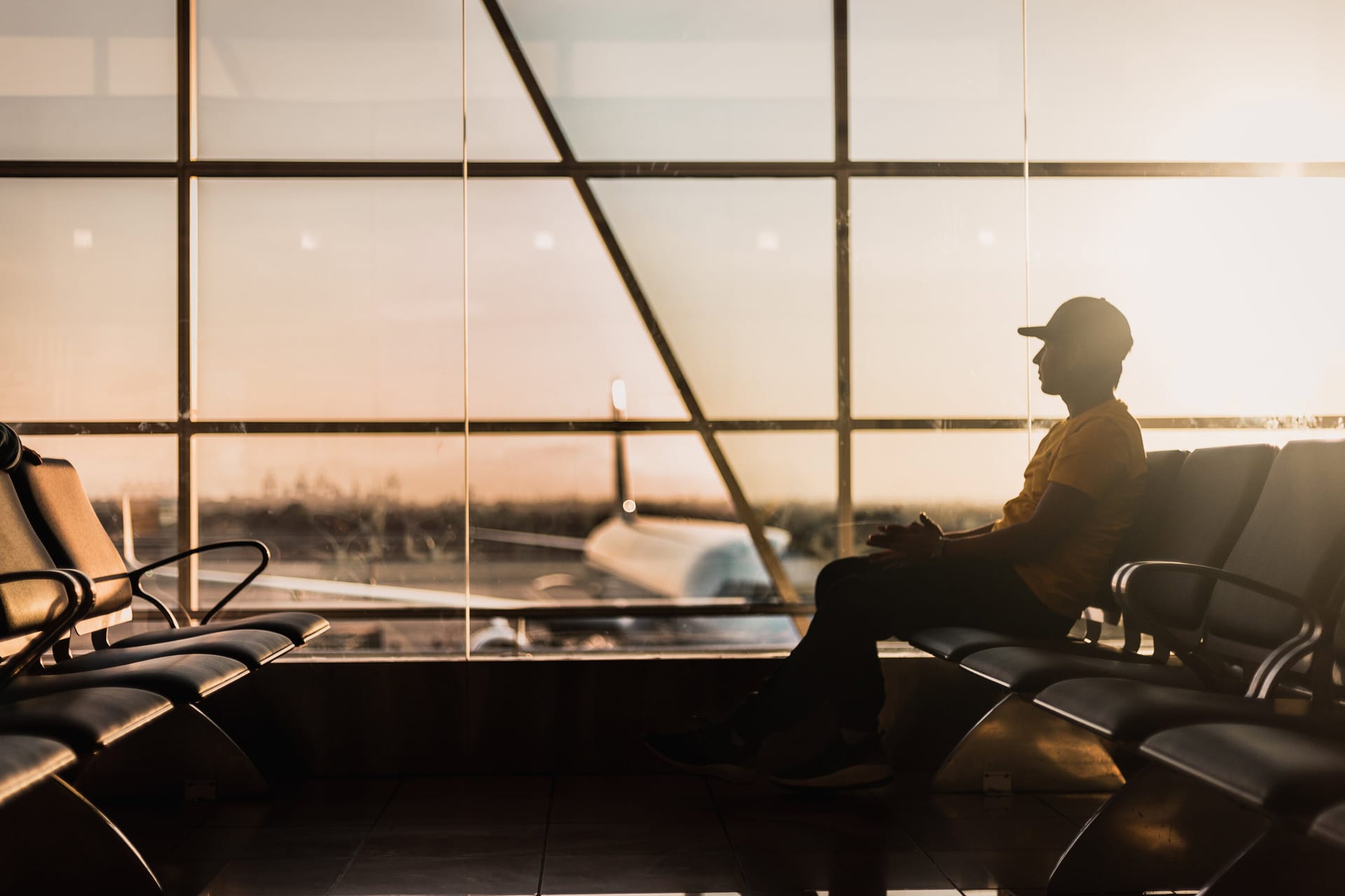 Silhouette of person waiting in airport terminal at sunset, with aircraft visible through large windows.