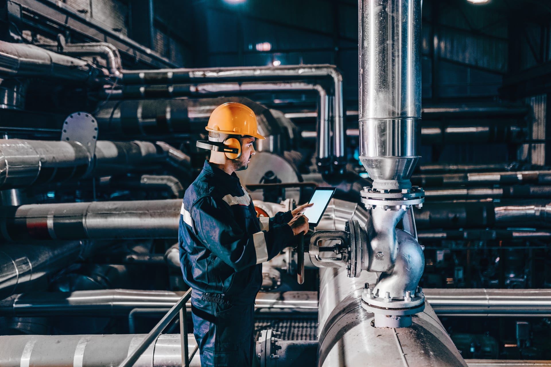 Industrial worker in orange hard hat and ear protection using tablet to monitor equipment in a factory pipeline system.