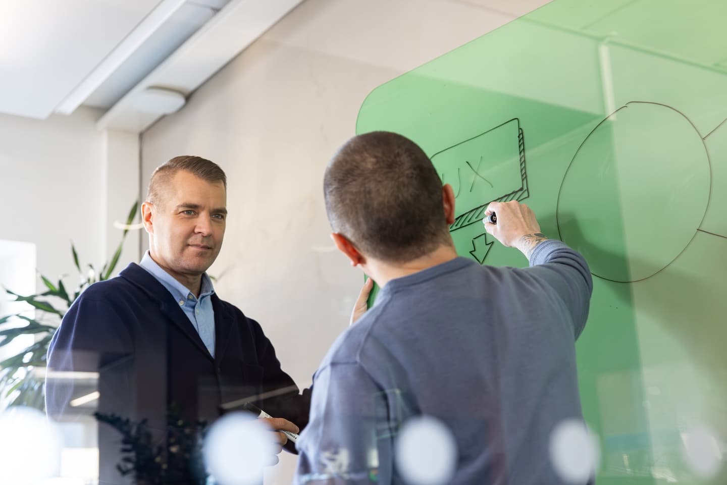 Two colleagues in an office, one drawing a diagram on a green glass board while the other watches attentively.