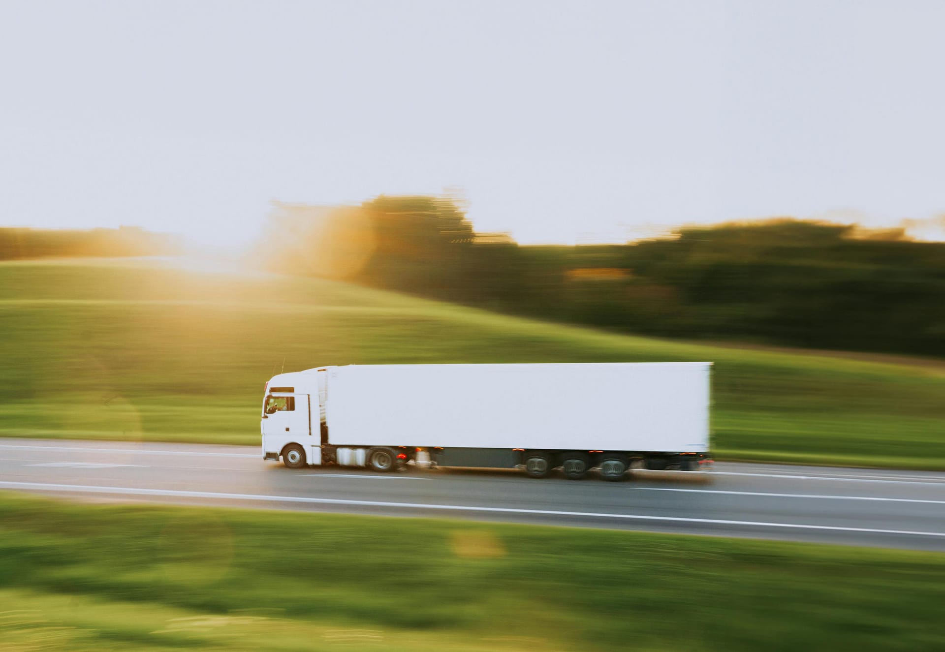 A white articulated lorry speeding along a road surrounded by green fields and trees, with warm golden sunlight in the background.