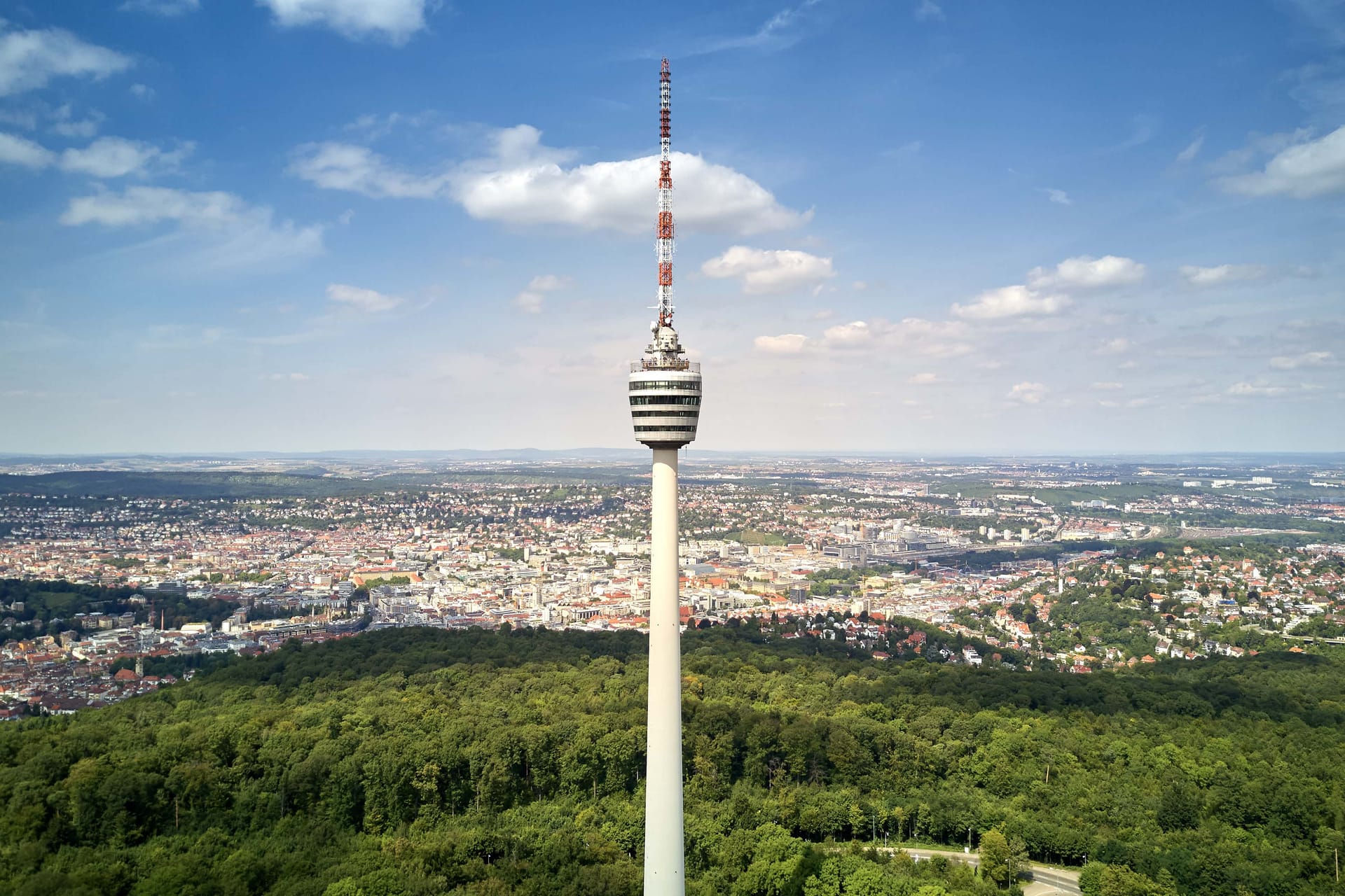 Tall television tower with striped observation deck rising above forest with cityscape view under blue sky with clouds.