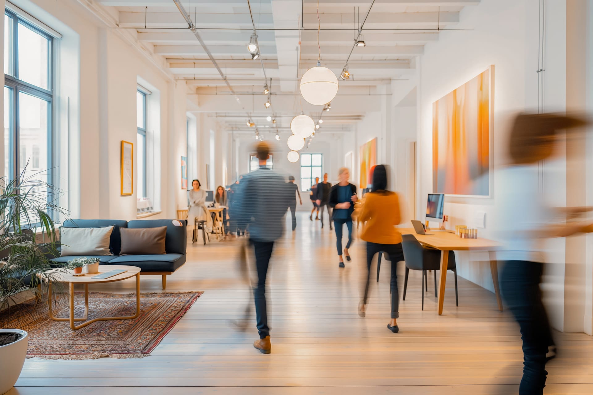 Bright modern coworking space with blurred people walking, globe pendant lights, wooden floors, and orange artwork on walls.
