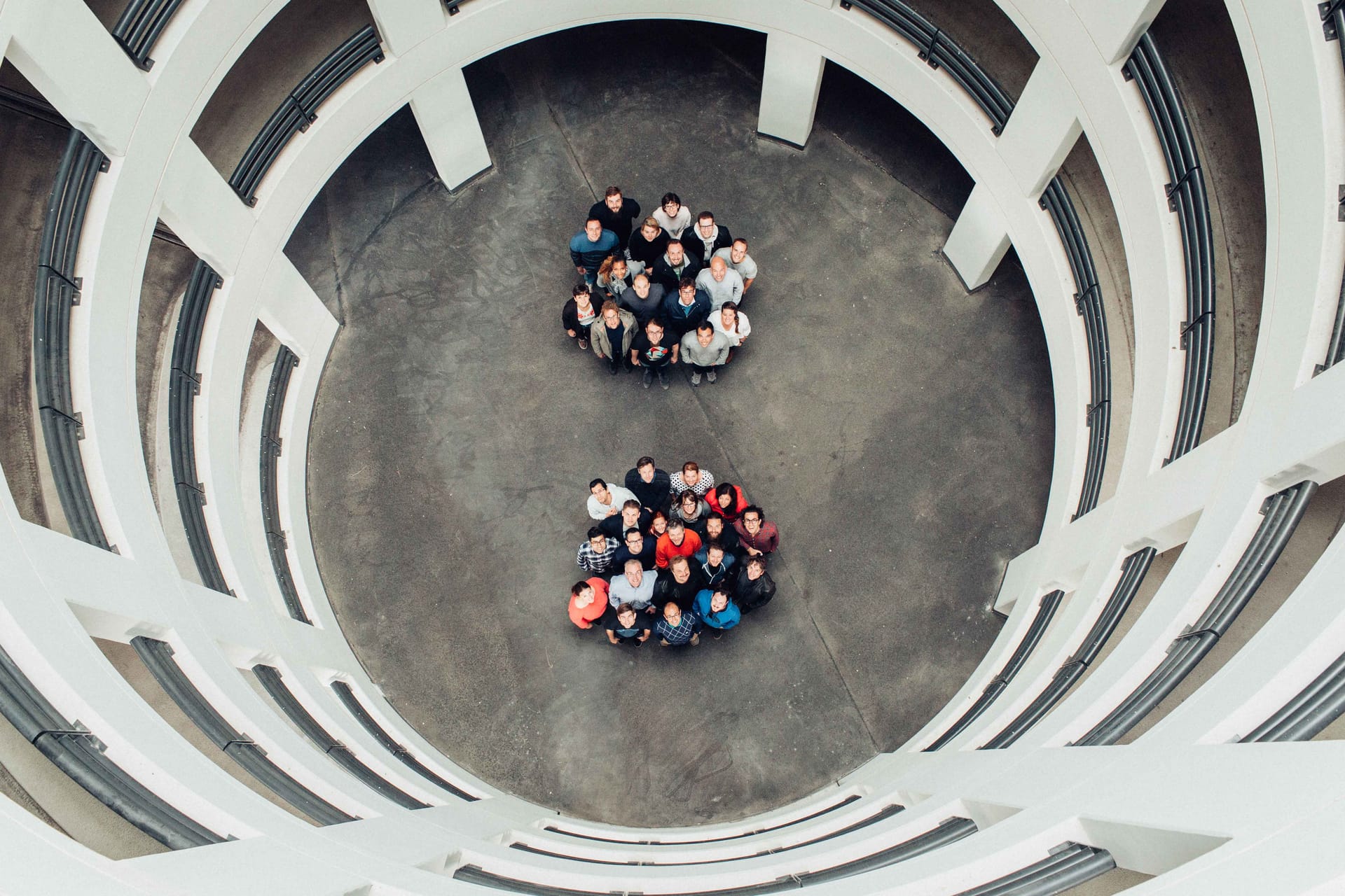 Aerial view of two groups of people standing in circular formations on a concrete floor surrounded by curved white architecture.