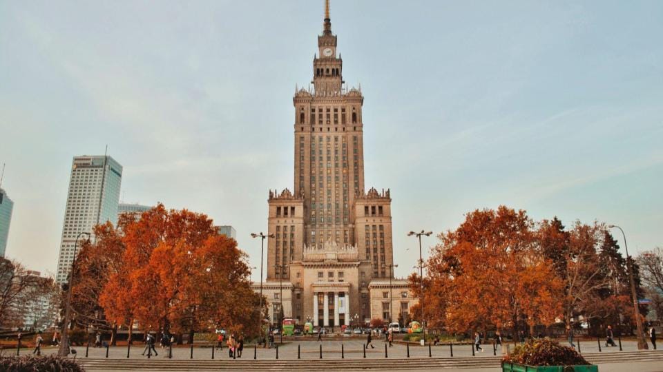 Palace of Culture and Science in Warsaw surrounded by autumn trees with vibrant orange foliage against a clear sky.