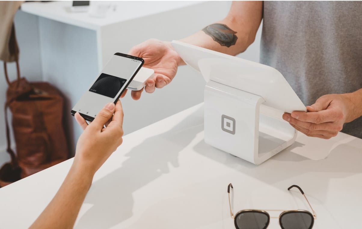 Person making contactless payment with smartphone at a Square payment terminal on white counter, sunglasses visible.
