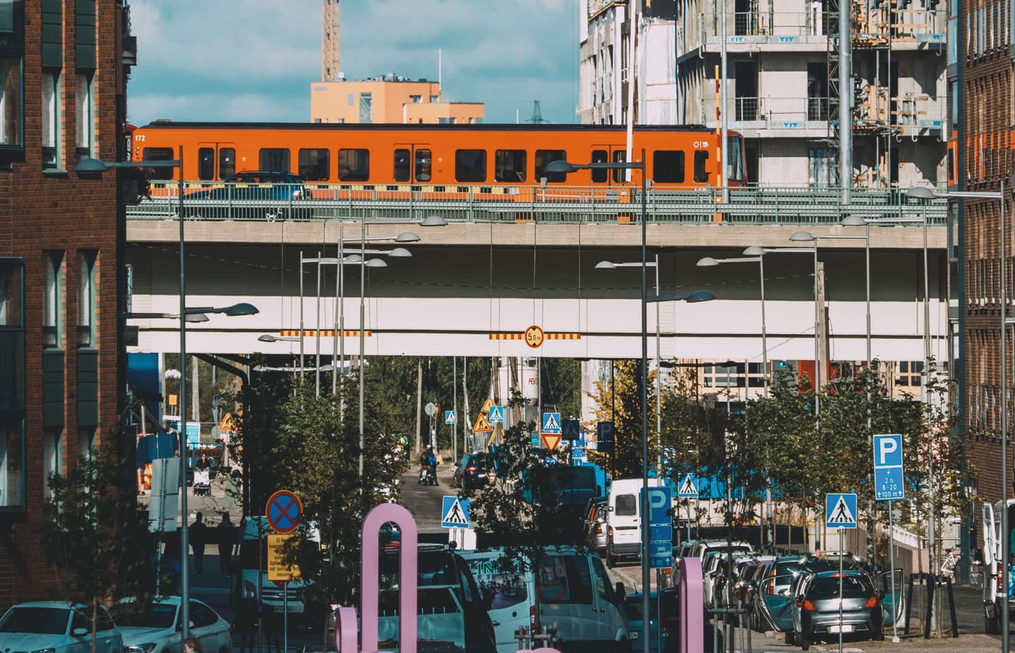 Bright orange train crossing an elevated railway bridge in an urban setting with traffic and road signs below.