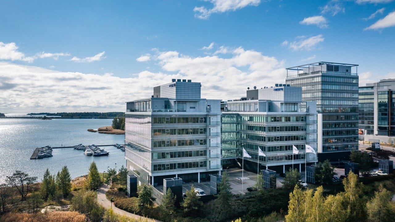 Modern glass office building of Neste overlooking a marina with boats, surrounded by trees and water under a blue sky with clouds.