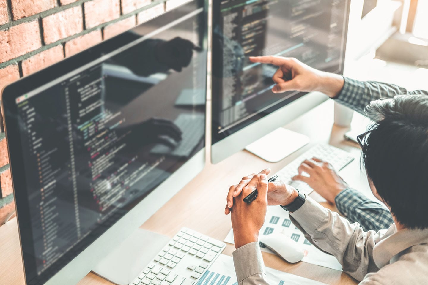Programmer pointing at code on dual monitors while collaborating with colleague in office with brick wall background.