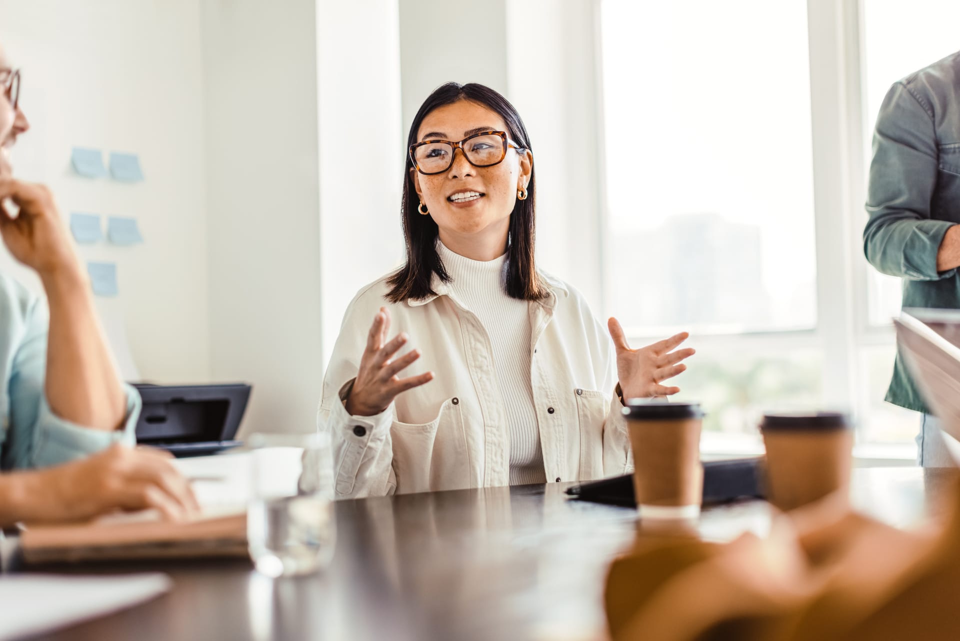 Woman in glasses and white outfit speaking expressively during a meeting with coffee cups on the table in a bright office.