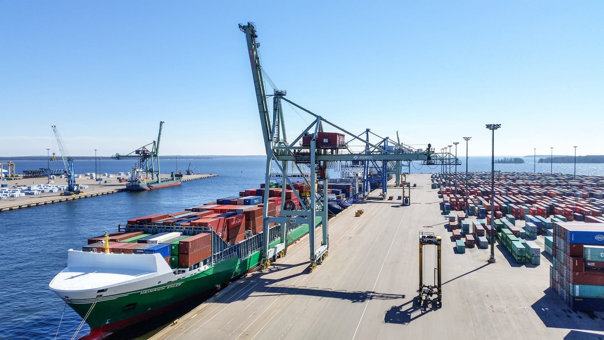 Busy shipping port with cargo vessel loaded with colourful containers, large cranes and stacked containers on the dock.