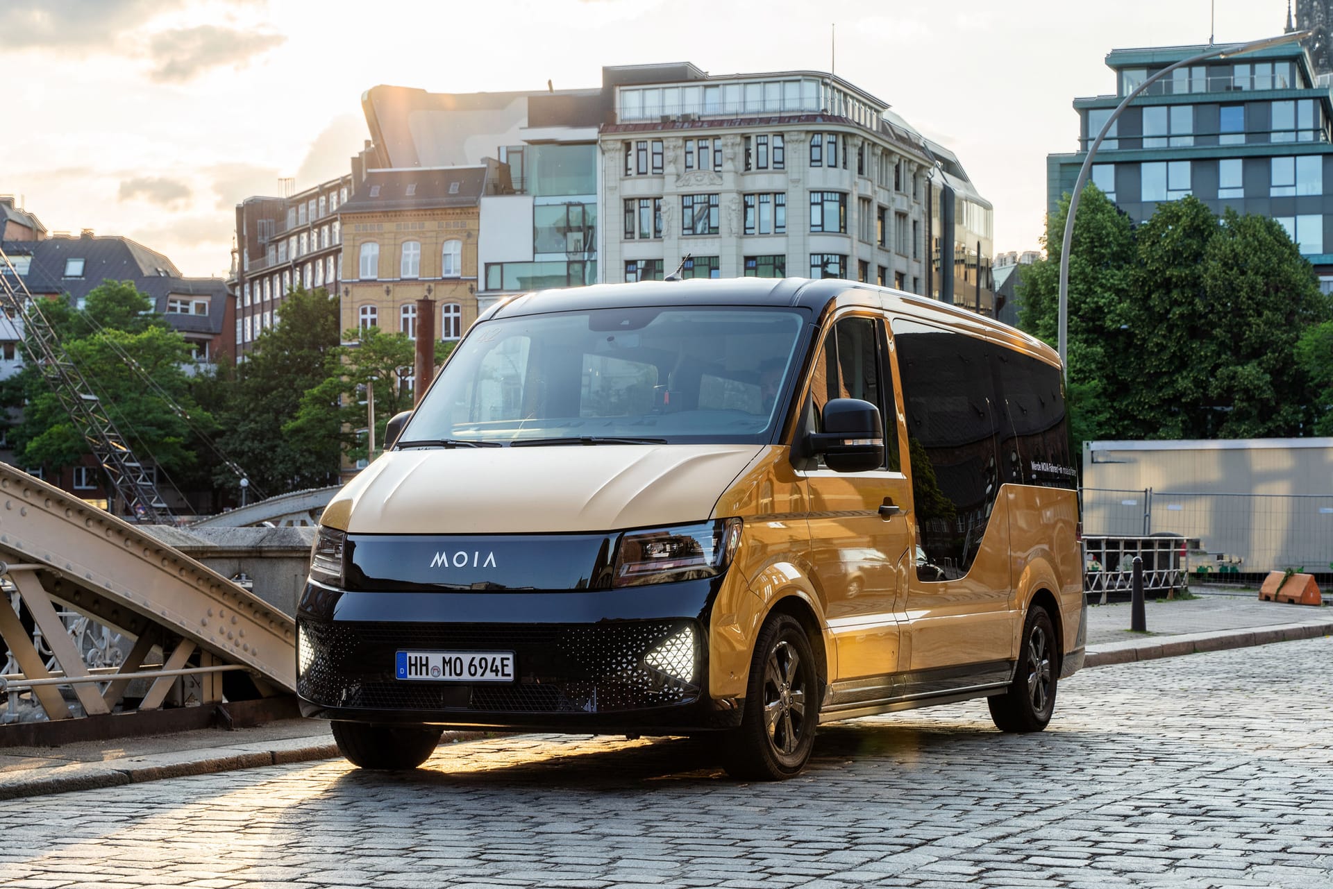 Gold MOIA electric van parked on cobblestone street in urban setting with historic buildings and sunset lighting.